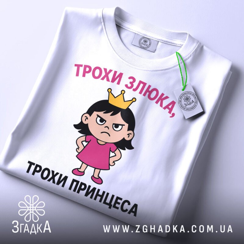 Футболка трохи злюка, біла, зображена з верхнього ракурсу, фон світло-сірий, принт з дівчинкою в короні та написом 'ТРОХИ ЗЛЮКА, ТРОХИ ПРИНЦЕСА', на футболці є етикетка з зеленим шнурком.