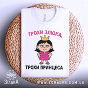 Футболка трохи злюка, біла, зображена зверху на плетеній кошику, фон світло-сірий, принт з дівчинкою в короні та написом 'ТРОХИ ЗЛЮКА, ТРОХИ ПРИНЦЕСА'.
