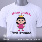 Футболка трохи злюка, біла, зображена з верхнього ракурсу, фон світло-сірий, принт з дівчинкою в короні та написом 'ТРОХИ ЗЛЮКА, ТРОХИ ПРИНЦЕСА', на футболці є етикетка з зеленим шнурком.