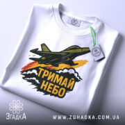 Футболка тримай небо. Біла футболка, складена на столі, з принтом літака, що летить, та написом 'ТРИМАЙ НЕБО'. Фон світлий, футболка має етикетку з зеленим шнурком, деталі принта чіткі та кольорові.
