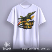 Футболка тримай небо. Біла футболка, підвішена на вішалці, з принтом літака, що летить, та написом 'ТРИМАЙ НЕБО'. Фон світло-сірий, принт яскравий з червоними та жовтими елементами.