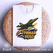 Футболка тримай небо. Біла футболка, складена на плетеній кошику, з принтом літака, що летить, та написом 'ТРИМАЙ НЕБО'. Фон світлий, деталі принта яскраві, з акцентами жовтого та червоного кольорів.