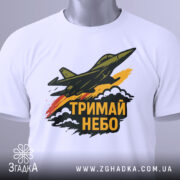 Футболка тримай небо. Біла футболка, складена на столі, з принтом літака, що летить, та написом 'ТРИМАЙ НЕБО'. Фон світлий, футболка має етикетку з зеленим шнурком, деталі принта чіткі та кольорові.
