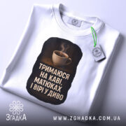 Футболка тримаюся на каві, складена на столі, біла, з етикеткою, фон світло-сірий, принт з чашкою кави та текстом на чорному фоні.