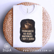 Футболка тримаюся на каві, складена на столі, біла, з етикеткою, фон світло-сірий, принт з чашкою кави та текстом на чорному фоні.