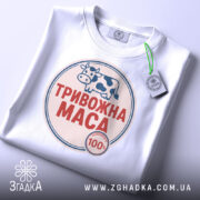 Футболка тривожна маса, біла, складена на столі, фон світло-сірий, принт з коровою та написом "ТРИВОЖНА МАСА" червоного кольору, з етикеткою на зеленому шнурку.