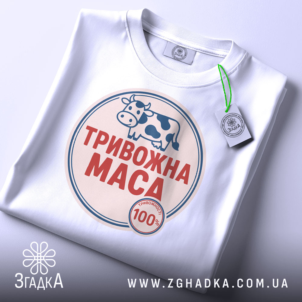 Футболка тривожна маса, біла, складена на столі, фон світло-сірий, принт з коровою та написом "ТРИВОЖНА МАСА" червоного кольору, з етикеткою на зеленому шнурку.