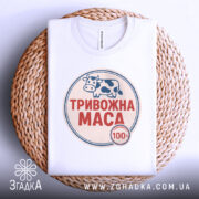 Футболка тривожна маса, біла, складена на плетеній кошику, фон світло-сірий, принт з коровою та написом "ТРИВОЖНА МАСА" червоного кольору.