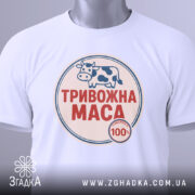 Футболка тривожна маса, біла, складена на столі, фон світло-сірий, принт з коровою та написом "ТРИВОЖНА МАСА" червоного кольору, з етикеткою на зеленому шнурку.