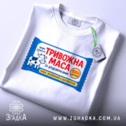 Футболка тривожна маса білого кольору, зображена зверху, фон світлий, на футболці яскравий принт з коровою та текстом "ТРИВОЖНА МАСА", на комірці видна етикетка.