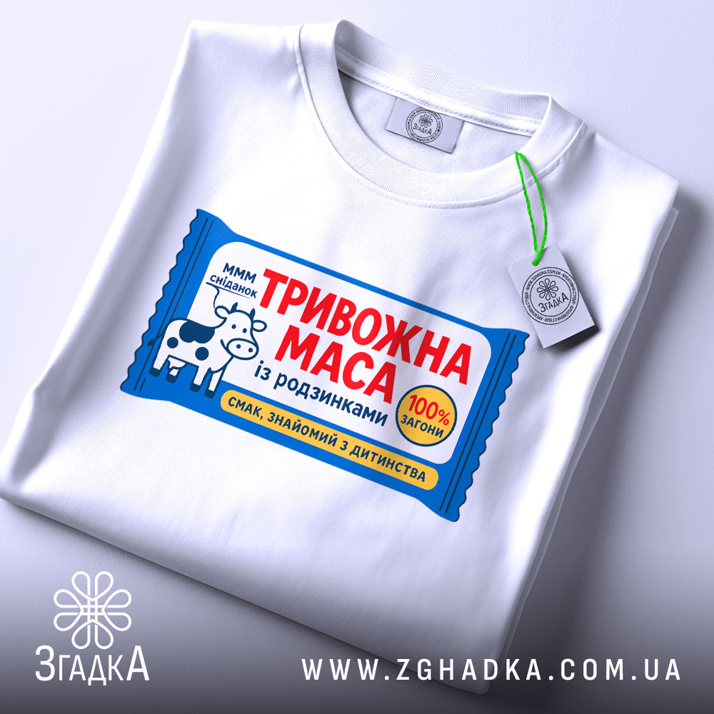 Футболка тривожна маса білого кольору, зображена зверху, фон світлий, на футболці яскравий принт з коровою та текстом "ТРИВОЖНА МАСА", на комірці видна етикетка.