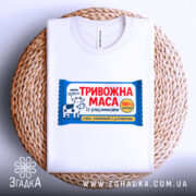 Футболка тривожна маса білого кольору, розкладена на плетеній основі, фон нейтральний, принт яскравий, з коровою та текстом "ТРИВОЖНА МАСА".