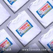 Футболка тривожна маса білого кольору, зображена з верхнього ракурсу, фон сірий, кілька футболок з принтом "ТРИВОЖНА МАСА" розкладені поруч.