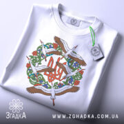 Футболка Тризуб Лелеки, біла, складена на поверхні, фон світло-сірий, принт з тризубом та лелеками, на футболці є етикетка з зеленим шнурком.