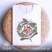Футболка Тризуб Лелеки, біла, складена на плетеній кошику, фон світло-сірий, принт з тризубом та лелеками, видно етикетку з написом.