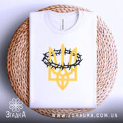 Футболка тризуб терни білого кольору, складена на круглій кошику з плетеного матеріалу, фон світло-сірий, принт у вигляді тризуба жовтого кольору з чорними тернами, видно етикетку на комірці.