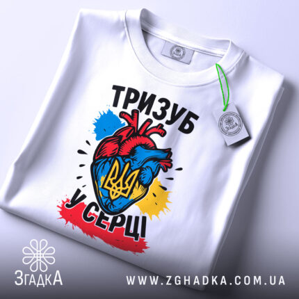 Футболка тризуб у серці, біла, зображена зверху, фон світло-сірий, принт з серцем у кольорах України та тризубом, на футболці є етикетка з зеленим шнурком.