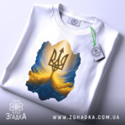 Футболка тризуб золото, біла, зображена зверху, на столі, фон світлий, принт з тризубом у золотих та блакитних тонах, з етикеткою зеленого кольору.