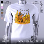 Футболка це добре, біла, з принтом, що зображує собаку в капелюсі, що сидить на стільці біля столу з чашкою, оточений полум'ям. Фон нейтральний, акцент на яскравих кольорах принта.