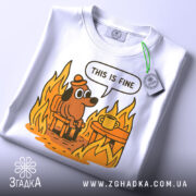 Футболка це добре, біла, складена, з принтом собаки в капелюсі, що сидить на стільці біля столу з чашкою, оточений полум'ям. Яскраві кольори принта на світлому фоні, видно етикетку.