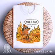 Футболка це добре, біла, складена на плетеній основі, з принтом собаки в капелюсі, що сидить на стільці біля столу з чашкою, оточений полум'ям. Яскраві кольори принта контрастують з текстурою основи.