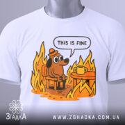 Футболка це добре, біла, складена, з принтом собаки в капелюсі, що сидить на стільці біля столу з чашкою, оточений полум'ям. Яскраві кольори принта на світлому фоні, видно етикетку.