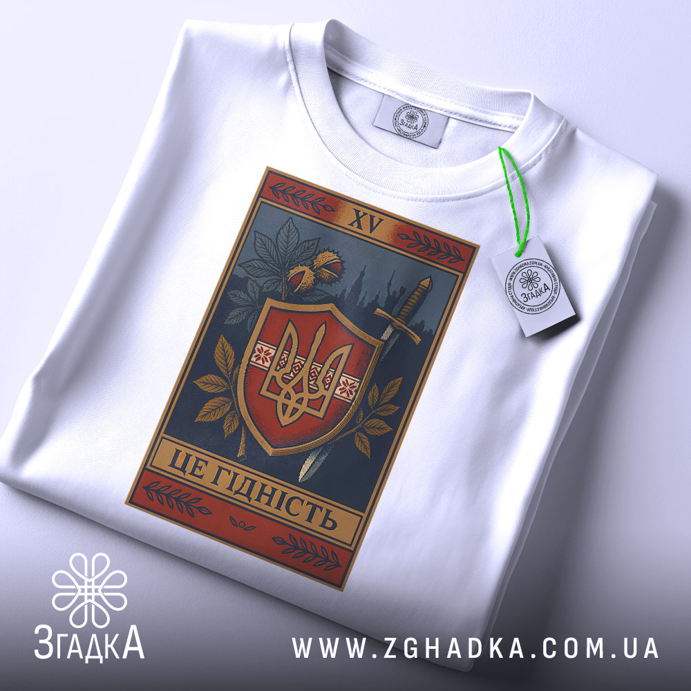 Футболка це гідність. Біла футболка, складена на поверхні, з видимим принтом герба України. На фоні світло-сірого градієнту, з зеленим ярликом, що підкреслює деталі дизайну. Принт містить рослинні елементи та напис 'ЦЕ ГІДНІСТЬ'.