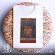 Футболка це гідність. Біла футболка, складена на плетеній кошику, з видимим принтом герба України. Фон світлий, а принт містить рослинні елементи та напис 'ЦЕ ГІДНІСТЬ'.