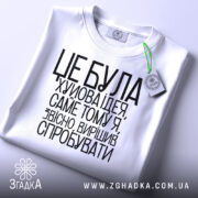 Футболка це погана ідея. Біла футболка, складена на столі, з чорним принтом. На футболці є зелений ярлик. Фон світло-сірий, текст добре читається, розташований по центру.