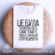 Футболка це погана ідея. Біла футболка, складена на плетеній основі, з чорним принтом. Фон світло-сірий, текст добре читається, розташований по центру.