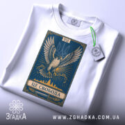 Футболка це свобода, біла футболка, складена на поверхні, принт з орлом на синьому фоні, ракурс зверху, фон світлий, на футболці є етикетка з зеленим шнурком.