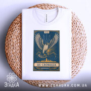 Футболка це свобода, біла футболка, складена на плетеній основі, принт з орлом на синьому фоні, ракурс зверху, фон світлий, деталі принта включають напис 'ЦЕ СВОБОДА' та герб України.