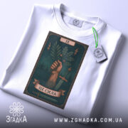 Футболка це сила. Біла футболка, складена на поверхні, з видимим принтом, зображена з верхнього ракурсу. Принт з рукою, що тримає гілку, на фоні темно-зеленого кольору, з елементами блискавок і мечів, з етикеткою, що звисає.