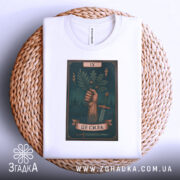 Футболка це сила. Біла футболка, складена на плетеній кошику, зображена з верхнього ракурсу. Принт з рукою, що тримає гілку, на фоні темно-зеленого кольору, з елементами блискавок і мечів, з видимим ярликом.