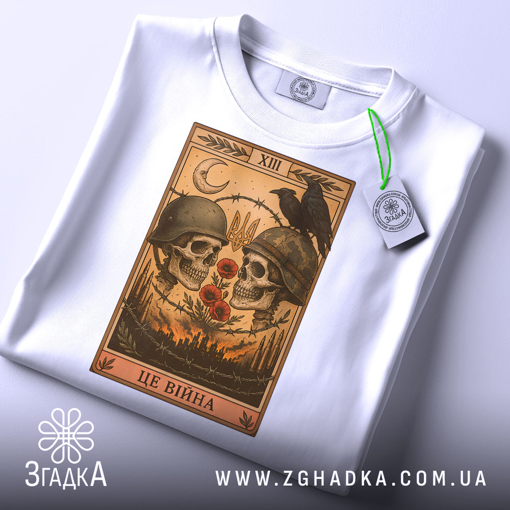 Футболка це війна. Біла футболка, складена на поверхні, з принтом черепів у шоломах, червоними квітами та чорним вороном. Вид з верхнього ракурсу, на футболці є етикетка з зеленим шнурком, фон світлий.