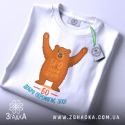 Футболка твори добро. Біла футболка, складена на столі, ведмідь з піднятими руками, текст "БРО ТВОРИ ДОБРО" та "БО ДОБРО ПЕРЕМАГАЄ ЗЛО!" на принті, зелена бирка з логотипом, фон світлий, акцент на принті та бирці.