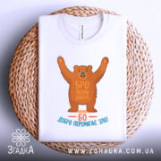 Футболка твори добро. Біла футболка, складена на плетеному кошику, ведмідь з піднятими руками, текст "БРО ТВОРИ ДОБРО" та "БО ДОБРО ПЕРЕМАГАЄ ЗЛО!" на принті, фон натуральний, акцент на принті та текстурі кошика.