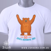 Футболка твори добро. Біла футболка, складена на столі, ведмідь з піднятими руками, текст "БРО ТВОРИ ДОБРО" та "БО ДОБРО ПЕРЕМАГАЄ ЗЛО!" на принті, зелена бирка з логотипом, фон світлий, акцент на принті та бирці.