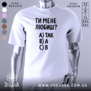 Футболка ти мене любиш? Біла футболка з чорним принтом, зображена з фронтального ракурсу на манекені. На фоні світло-сірий градієнт. Принт містить текст: 'ТИ МЕНЕ ЛЮБИШ?' та варіанти відповідей A) ТАК, B) А, C) В.
