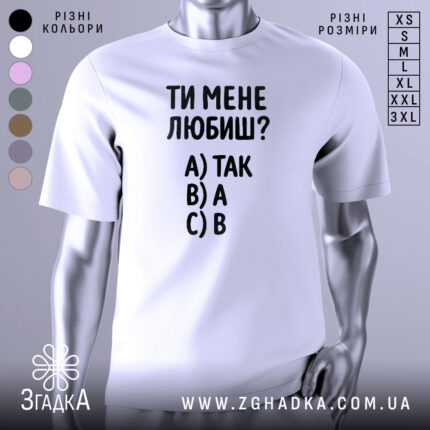 Футболка ти мене любиш? Біла футболка з чорним принтом, зображена з фронтального ракурсу на манекені. На фоні світло-сірий градієнт. Принт містить текст: 'ТИ МЕНЕ ЛЮБИШ?' та варіанти відповідей A) ТАК, B) А, C) В.