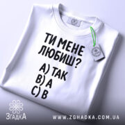 Футболка ти мене любиш? Біла футболка, складена на столі, з видимим етикеткою. Зображена з верхнього ракурсу. Фон світло-сірий. Принт містить текст: 'ТИ МЕНЕ ЛЮБИШ?' та варіанти відповідей A) ТАК, B) А, C) В.