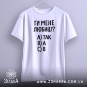 Футболка ти мене любиш? Біла футболка, підвішена на вішалці, зображена з фронтального ракурсу. Фон світло-сірий. Принт містить текст: 'ТИ МЕНЕ ЛЮБИШ?' та варіанти відповідей A) ТАК, B) А, C) В.