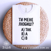 Футболка ти мене любиш? Біла футболка, складена на плетеному кошику, зображена з верхнього ракурсу. Фон світло-сірий. Принт містить текст: 'ТИ МЕНЕ ЛЮБИШ?' та варіанти відповідей A) ТАК, B) А, C) В.