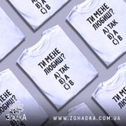 Футболка ти мене любиш? Біла футболка з чорним принтом, зображена з фронтального ракурсу на манекені. На фоні світло-сірий градієнт. Принт містить текст: 'ТИ МЕНЕ ЛЮБИШ?' та варіанти відповідей A) ТАК, B) А, C) В.