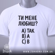 Футболка ти мене любиш? Біла футболка, складена на столі, з видимим етикеткою. Зображена з верхнього ракурсу. Фон світло-сірий. Принт містить текст: 'ТИ МЕНЕ ЛЮБИШ?' та варіанти відповідей A) ТАК, B) А, C) В.