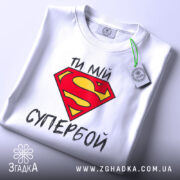 Футболка ти мій супербой, біла, з верхнього ракурсу, складена, з яскравим принтом у вигляді червоної літери S на жовтому фоні, з етикеткою на зеленому шнурку.