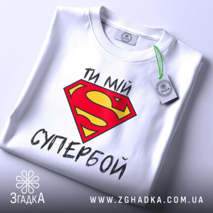 Футболка ти мій супербой, біла, з верхнього ракурсу, складена, з яскравим принтом у вигляді червоної літери S на жовтому фоні, з етикеткою на зеленому шнурку.