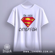 Футболка ти мій супербой, біла, з бокового ракурсу, підвішена на вішалці, з яскравим принтом у вигляді червоної літери S на жовтому фоні та текстом українською мовою.