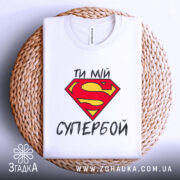 Футболка ти мій супербой, біла, з верхнього ракурсу, складена на плетеній основі, з яскравим принтом у вигляді червоної літери S на жовтому фоні та текстом українською мовою.