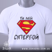 Футболка ти мій супербой, біла, з верхнього ракурсу, складена, з яскравим принтом у вигляді червоної літери S на жовтому фоні, з етикеткою на зеленому шнурку.